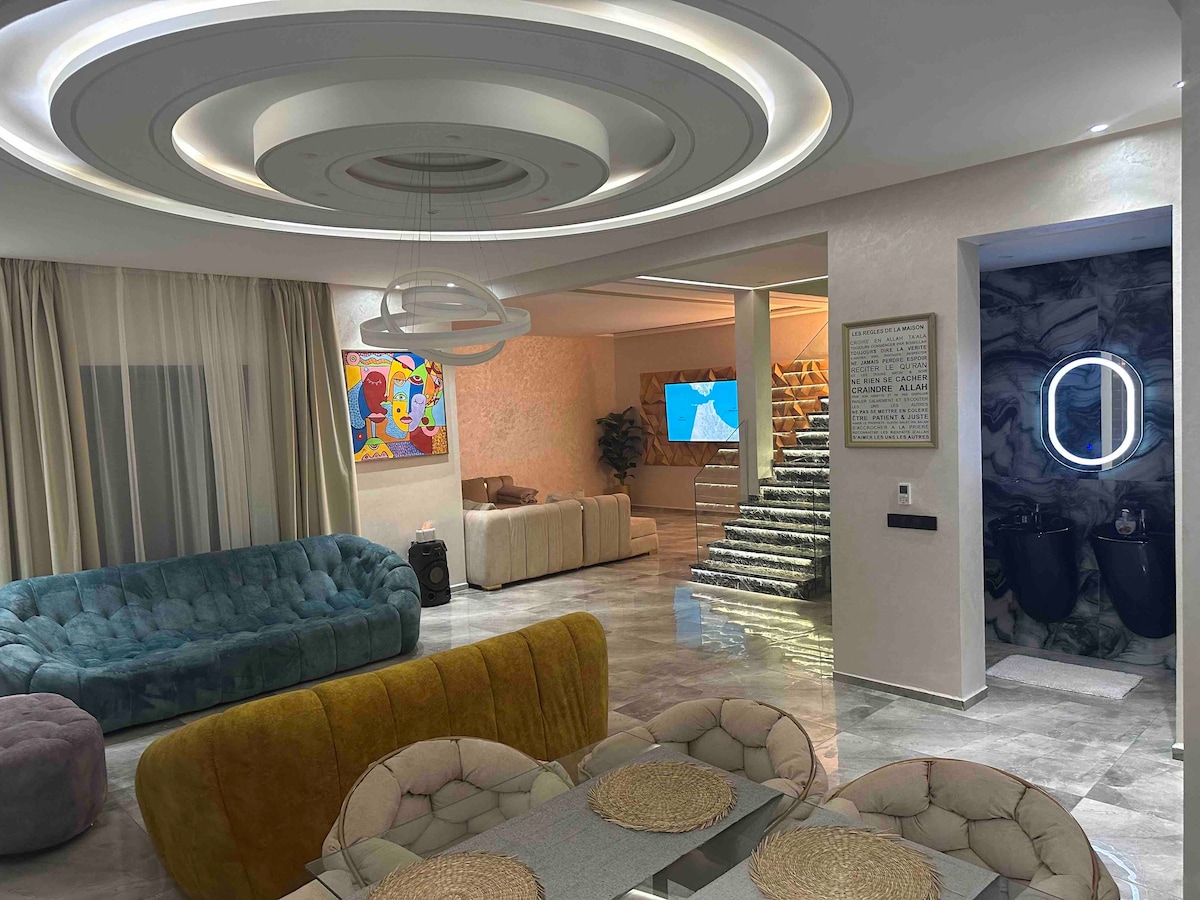 An inviting living area showcases a cozy blue sofa and a circular dining table set with natural-toned placemats. A colorful artwork hangs on the wall, and a softly illuminated staircase leads to the upper level. Light streams in through floor-to-ceiling windows, enhancing the spacious ambiance.