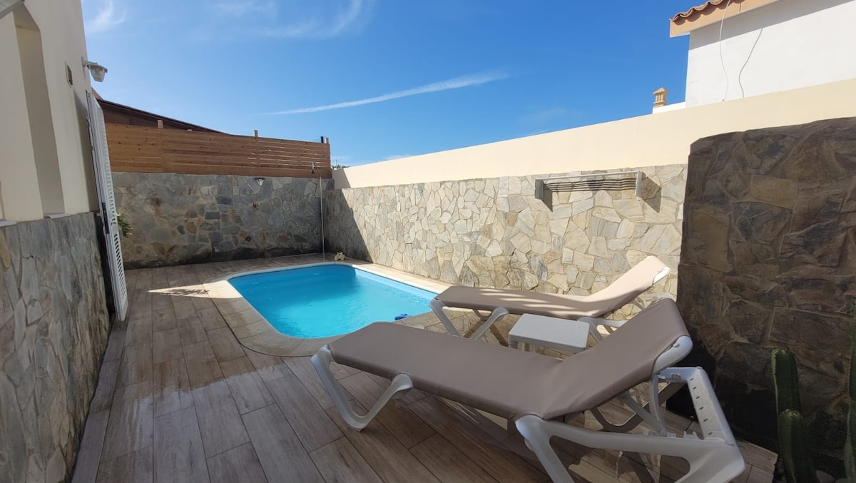 A private outdoor area features a clear pool surrounded by stone walls. Two loungers with light brown cushions are positioned beside the pool, offering a relaxing space. Bright blue skies are visible above, creating a serene setting.