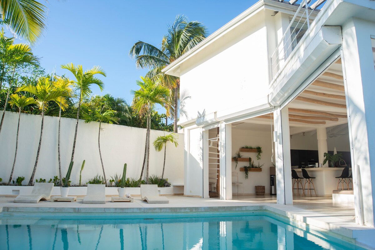 The villa features a refreshing pool surrounded by lounge chairs, with palm trees and cacti providing a tropical ambiance. Large glass doors open to an indoor space that connects seamlessly with the outdoor area, enhancing the villa's spacious feel.
