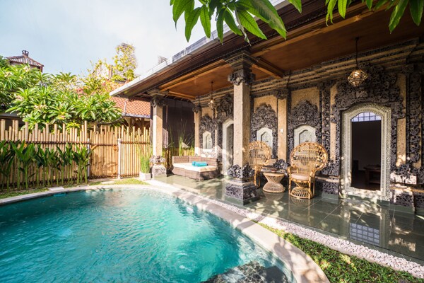 A private garden features a clear turquoise pool surrounded by lush greenery. Traditional Balinese architecture is displayed on the house's exterior, complemented by intricate stone carvings. Two rattan chairs are positioned near the pool, providing a serene spot for relaxation.