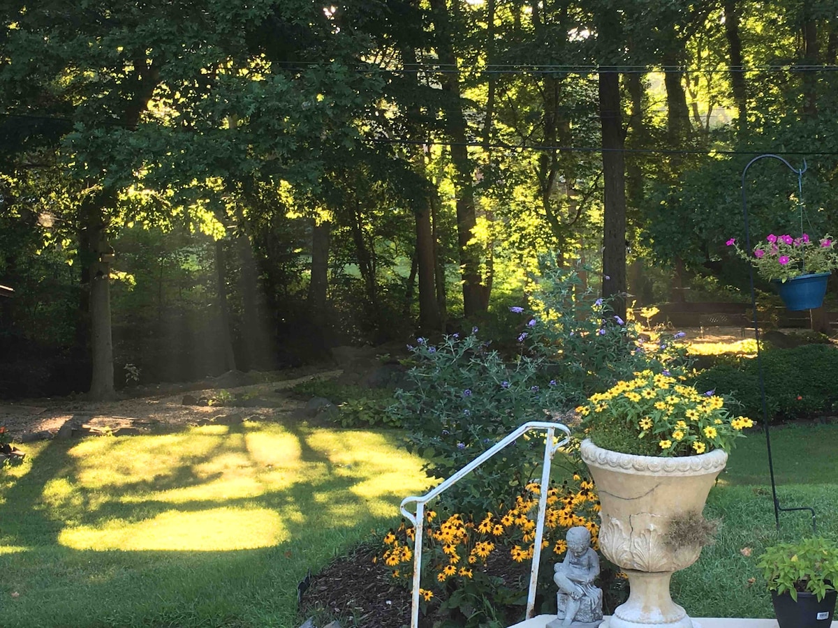 A well-maintained garden area is visible, featuring vibrant yellow flowers surrounding a decorative stone planter. Lush green trees create a serene backdrop, while soft sunlight filters through the leaves, casting gentle shadows on the grassy lawn.