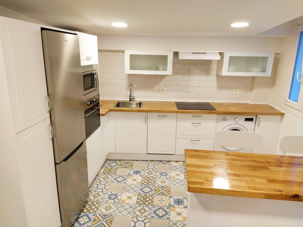 The kitchen features modern appliances, including a stainless steel refrigerator and oven. A sleek countertop with wooden accents provides supplementary space. Upper cabinets display glass fronts, offering visibility of stored items. Unique patterned tiles complete the look, adding an appealing touch to the cooking area.