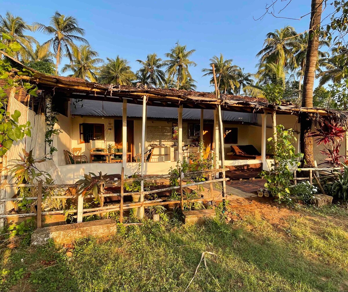 The exterior of a cozy house is framed by lush palm trees and greenery. A spacious porch with wooden supports is visible, offering a shaded area. Terracotta tiles and natural vegetation surround the entrance, creating a serene environment that welcomes relaxation.