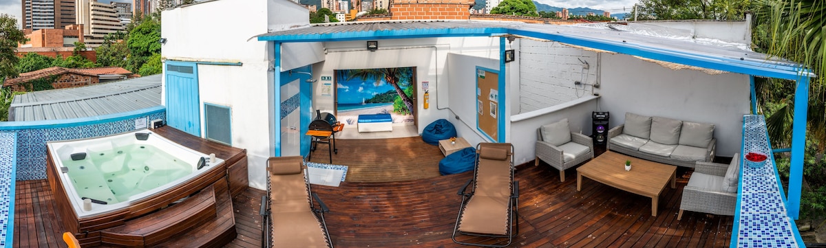 An outdoor terrace is depicted, featuring a spacious jacuzzi and comfortable lounge chairs. A wooden deck enhances the area, while various seating options, including bean bags and a modern couch, are arranged to create a welcoming space. Greenery is visible in the backdrop.