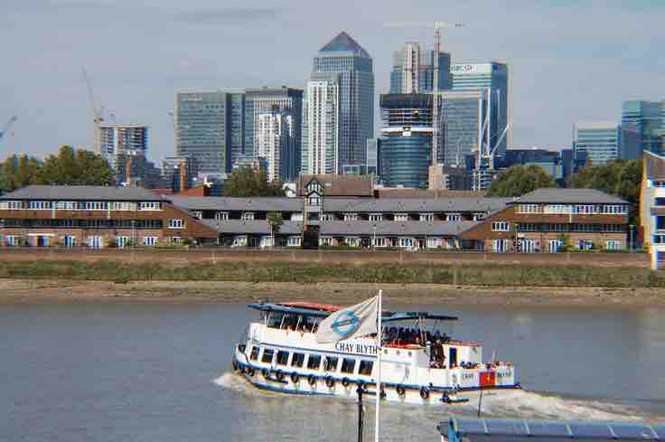 A riverboat travels along the Thames, offering views of the striking city skyline featuring modern high-rise buildings. The image captures the contrast between the boat and the urban landscape, with clear blue skies and calm water enhancing the scenery.