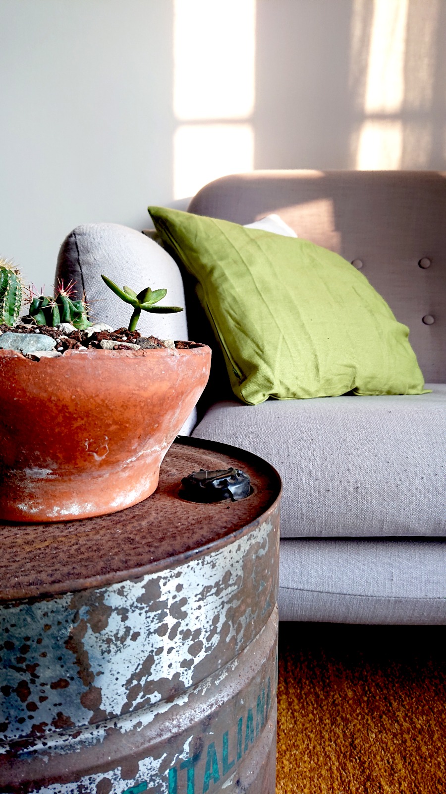 A cozy seating area is highlighted, featuring a light grey armchair with a green pillow. A rustic table made from a repurposed container sits nearby, adorned with a terracotta pot holding a small succulent. Natural light filters through a window, casting soft shadows.
