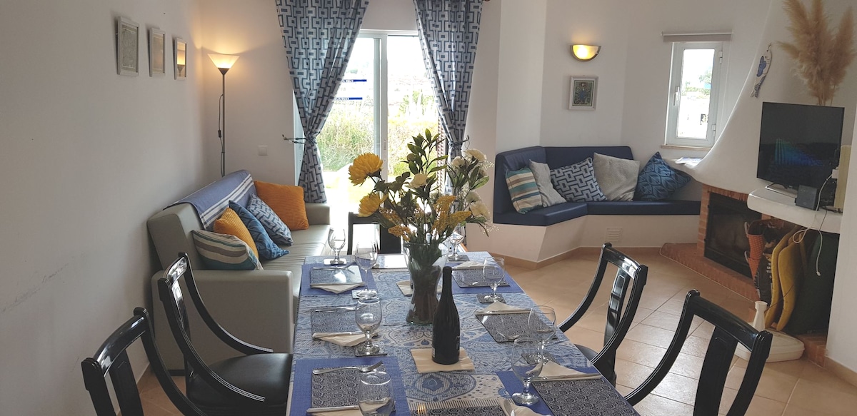 A dining area is set with a blue tablecloth, showcasing elegant tableware for six. Bright yellow flowers in a vase center the table. A cozy seating nook features cushions in various patterns, while large windows bring in natural light, enhancing the room's inviting feel.
