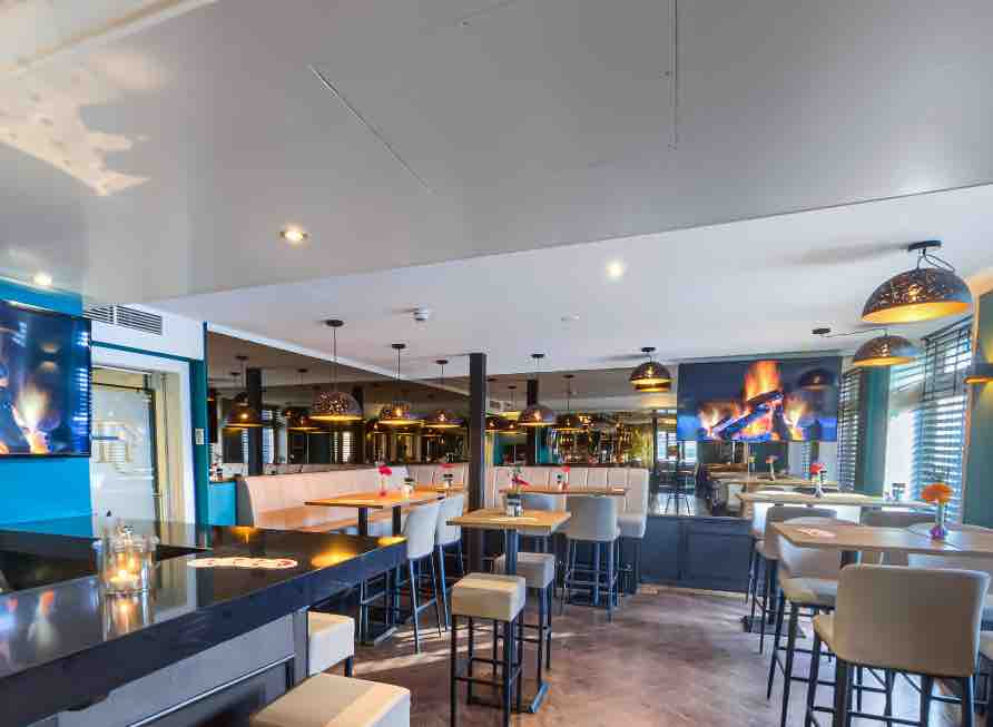A modern dining area is presented with a mix of high-top tables and cushioned seating. A fireplace mural adds warmth, complemented by subtle lighting fixtures. Large windows allow natural light to fill the space, enhancing the inviting ambiance of the restaurant.