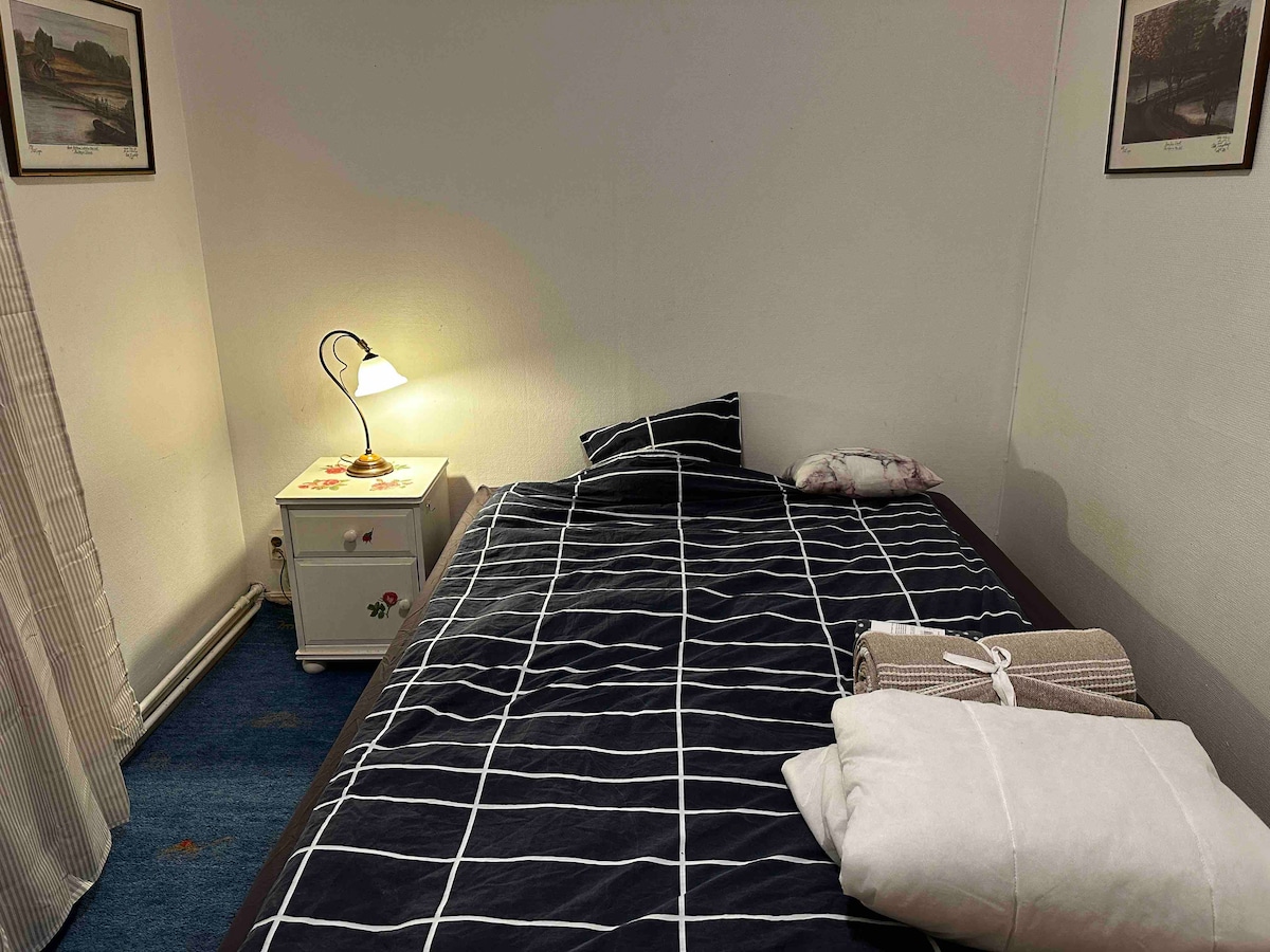A cozy sleeping area features a single bed adorned with a grid-patterned blanket. A bedside table with a lamp rests beside the bed, while additional linens are neatly rolled nearby. Soft lighting creates a serene atmosphere within the room.