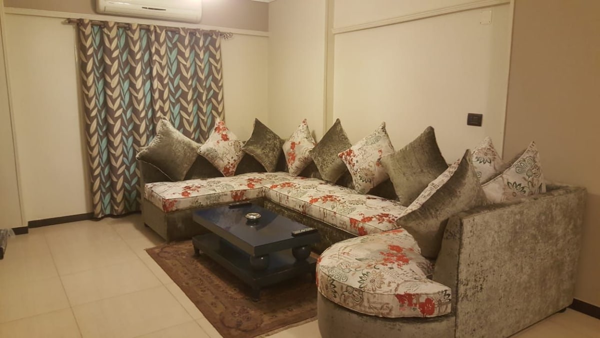 A spacious living area features a large sectional sofa adorned with floral and ornate cushions. A dark coffee table is positioned at the center, with a simple decorative item placed on it. Floor-to-ceiling curtains in a geometric pattern frame a nearby window.