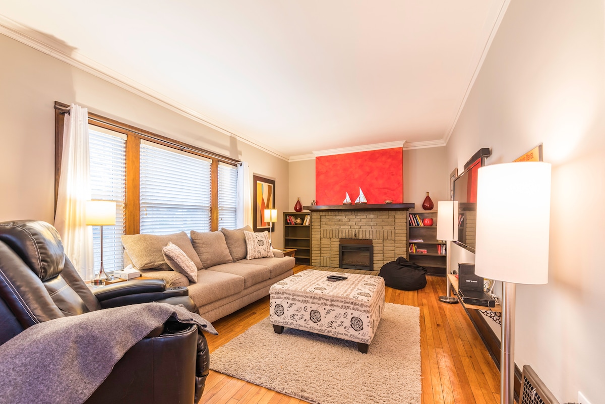 The living room features a spacious sectional sofa and a textured ottoman, complemented by a warm-hued accent wall. Natural light filters through the window with wooden blinds, and a cozy area rug adds comfort to the hardwood floor.