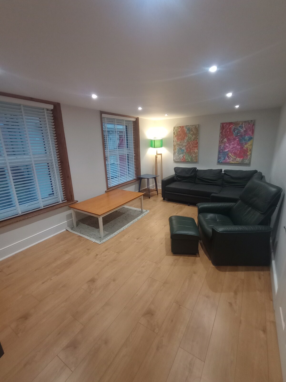 The lounge area features a dark leather sofa and an armchair paired with a wooden coffee table. Colorful artwork adorns the walls, and natural light filters through the slatted blinds. Modern lighting fixtures provide a bright ambiance throughout the space.