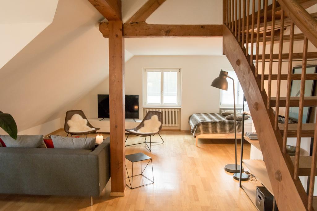 An open living space is highlighted by wooden beams and sleek furniture. Two chairs adorned with soft throws face a television. A spacious bed is set on a raised area, while a staircase leads to the upper level, enhancing the apartment's split-level design.