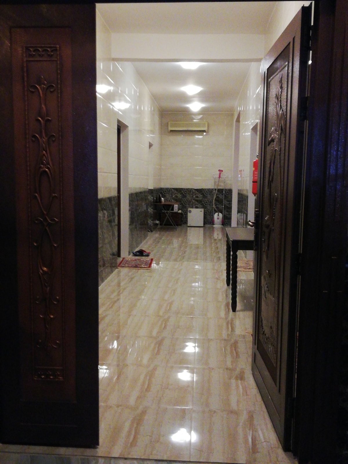 The entrance showcases dark wooden doors, opening to a tiled hallway with light-colored walls. Bright overhead lights illuminate the space, which features a minimalist design. A small table and chair are seen on the right, with a glimpse of additional furnishings in the background.