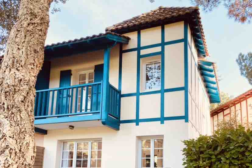 The exterior of a two-story villa features vibrant blue accents around the windows and balcony, nestled among trees. The rustic roof complements the charming architecture, while the well-maintained garden adds a touch of greenery to the setting.