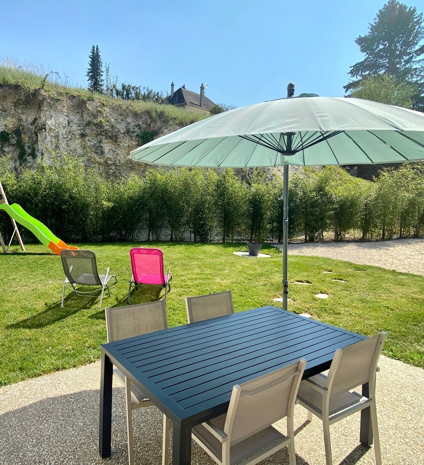 An outdoor space features a dining table with four chairs under a large umbrella. A slide is visible in the background, alongside two lounge chairs. Lush greenery surrounds the area, creating a private and peaceful environment for relaxation.