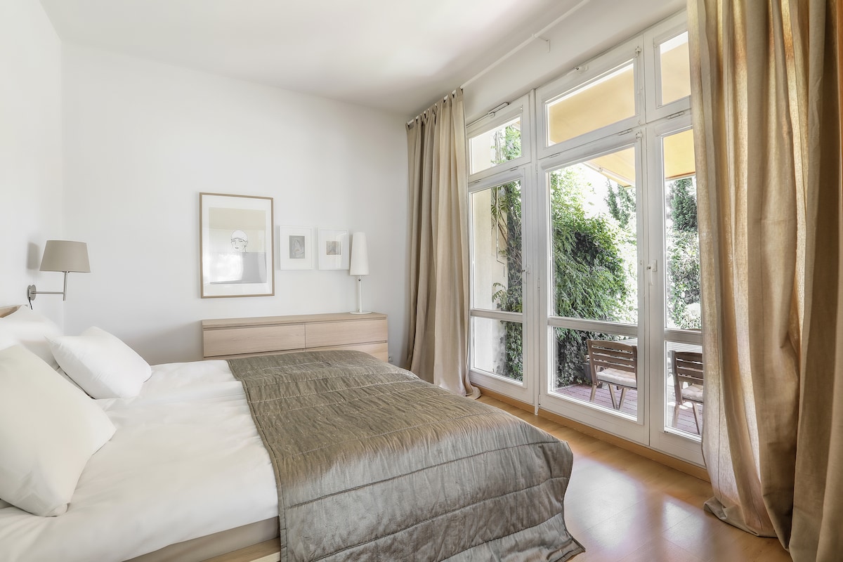 The spacious bedroom features large windows that overlook a well-maintained garden. Natural light fills the room, which includes a versatile bed and light-colored bedding. Elegant curtains frame the windows, complementing the neutral color palette of the space.