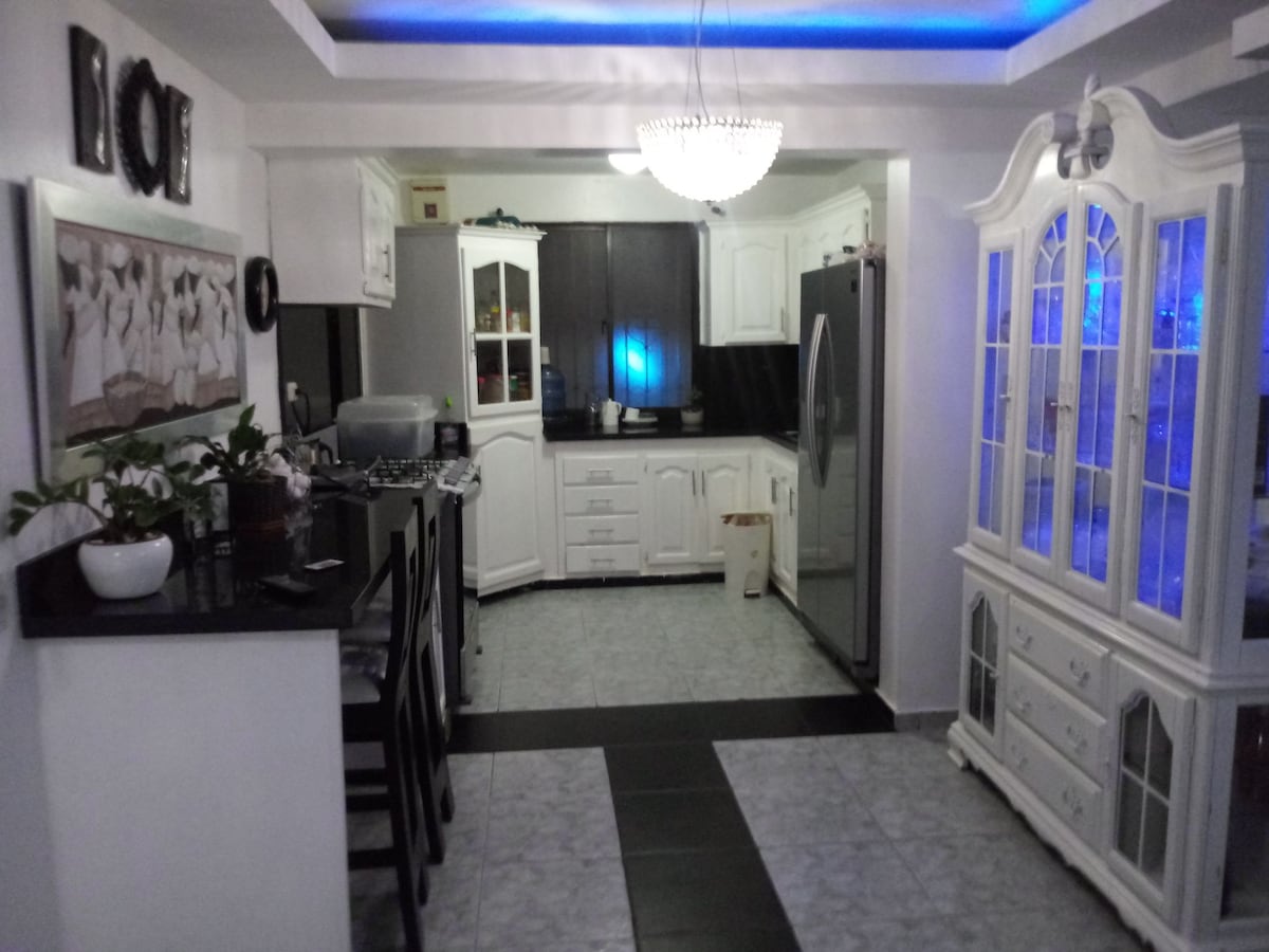 A bright kitchen features white cabinetry and modern appliances, including a large refrigerator. A central island with seating offers a gathering space, while a glass display cabinet adds a decorative element. Soft blue lighting enhances the ambiance in this contemporary kitchen.