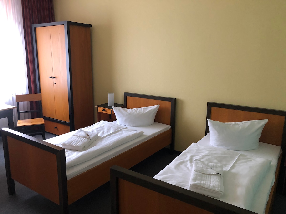 Two twin beds are arranged within a minimalistic room, featuring wooden frames and crisp white bedding. A wooden wardrobe and nightstand provide storage, while a simple chair is positioned nearby. Natural light filters through a window, casting a soft glow across the space.