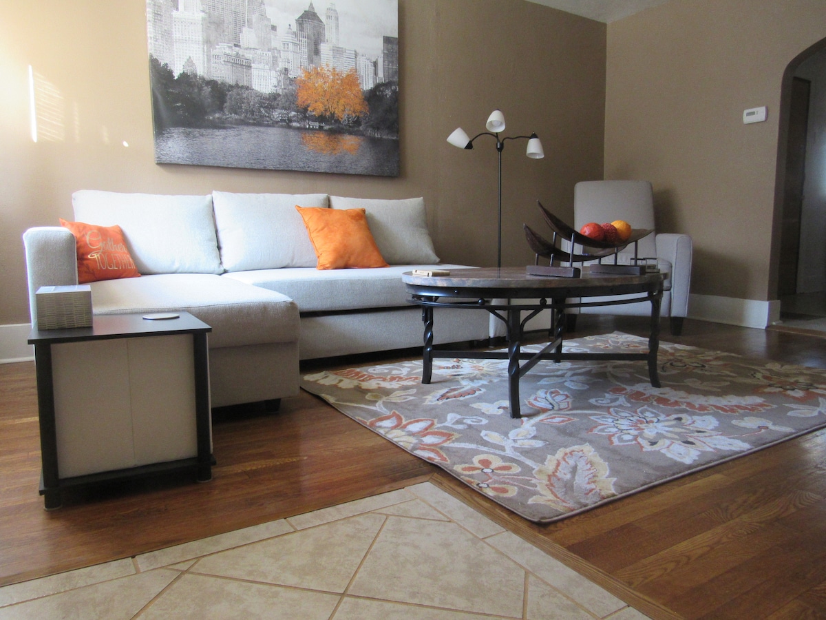 A living area features a light-colored sofa with orange accent pillows, accompanied by a round coffee table placed on a decorative area rug. Natural light filters in through the window, illuminating the space. Artwork depicting a cityscape adorns the wall.