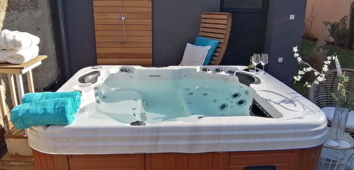 A hot tub, featuring an array of jets, is set against a neutral wall. Soft towels are neatly arranged nearby. Two glasses are placed on a small table, while a decorative vase with flowers adds a touch of elegance to the outdoor space.