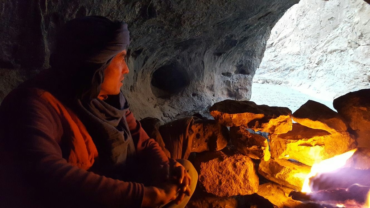 A warm fire burns within a stone circle, providing light and warmth in a cave setting. The rocky walls of the cave create a natural backdrop, with the entrance revealing a glimpse of the outdoors. The area is quiet and peaceful, inviting reflection.