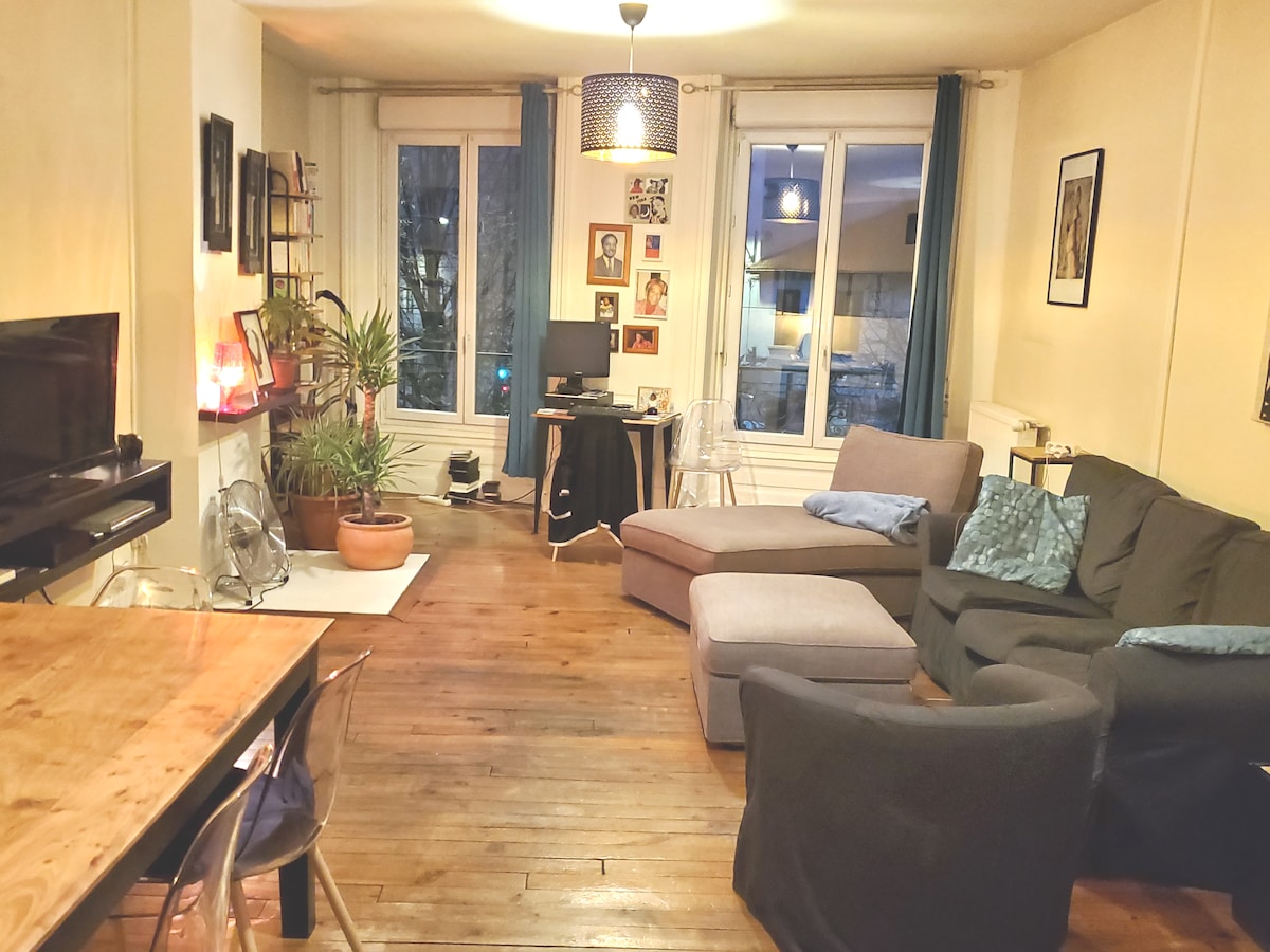 A well-lit living area features a grey sectional sofa and a matching armchair. A wooden dining table is set nearby. Large windows provide natural light and a view of the street. Decorative plants and framed photos add a personal touch to the space.