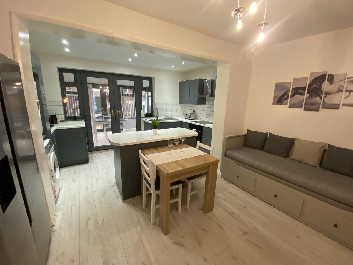 An open-plan kitchen and dining area is presented, showcasing a kitchen island at the center. A small wooden dining table is accompanied by white chairs. A designated seating area with a gray cushioned bench is visible, alongside modern kitchen appliances and doors leading to the garden.