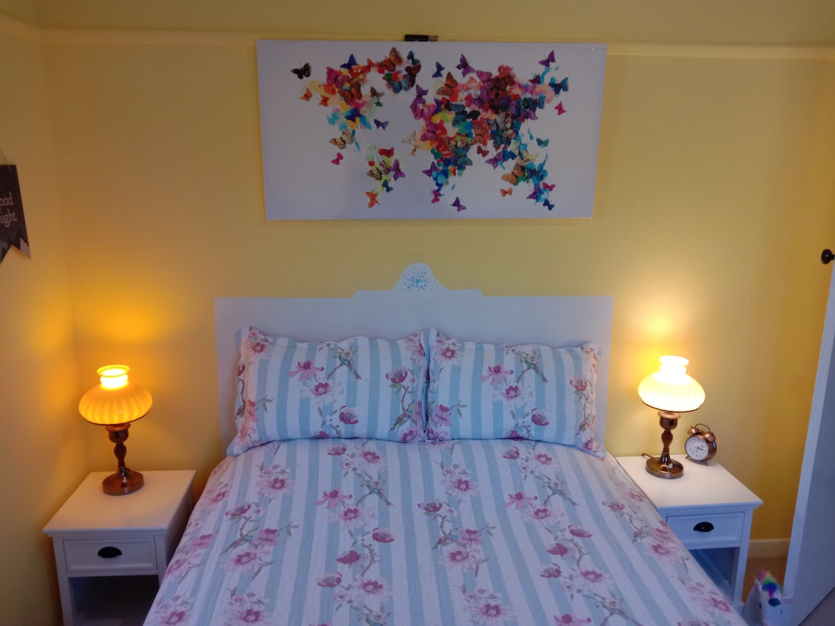 A cozy bedroom features a spacious bed with floral bedding, flanked by two lamps. A colorful butterfly wall art piece adds a cheerful touch above the bed, while a small clock sits on one bedside table against a bright yellow wall.