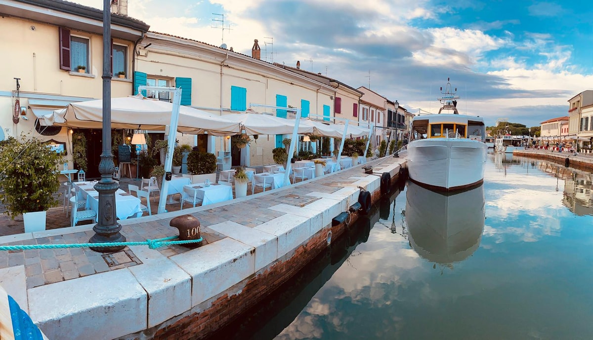 The serene waterfront scene features a canal lined with charming buildings, adorned with blue shutters. Dining tables and chairs are set up under canopies, creating a relaxed atmosphere. A boat is docked, reflecting softly on the calm water, while gentle clouds drift overhead.