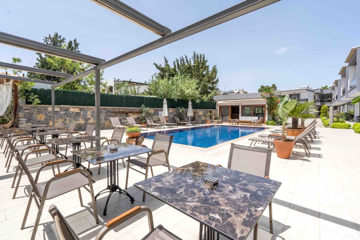 An outdoor pool area is presented, featuring a well-maintained swimming pool surrounded by comfortable lounge chairs. Several tables are positioned nearby, accompanied by lush greenery and umbrellas providing shade. The space is designed for relaxation with a serene ambiance.
