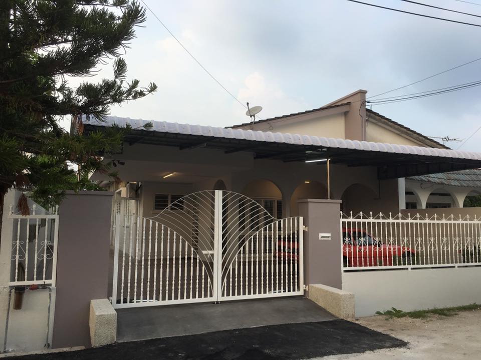 A semi-detached house is visible, featuring a white gate with leaf-patterned details. The driveway leads to a covered carport, accommodating a visible red vehicle. Arched windows and a roof with overhangs create a welcoming exterior, framed by greenery from nearby trees.