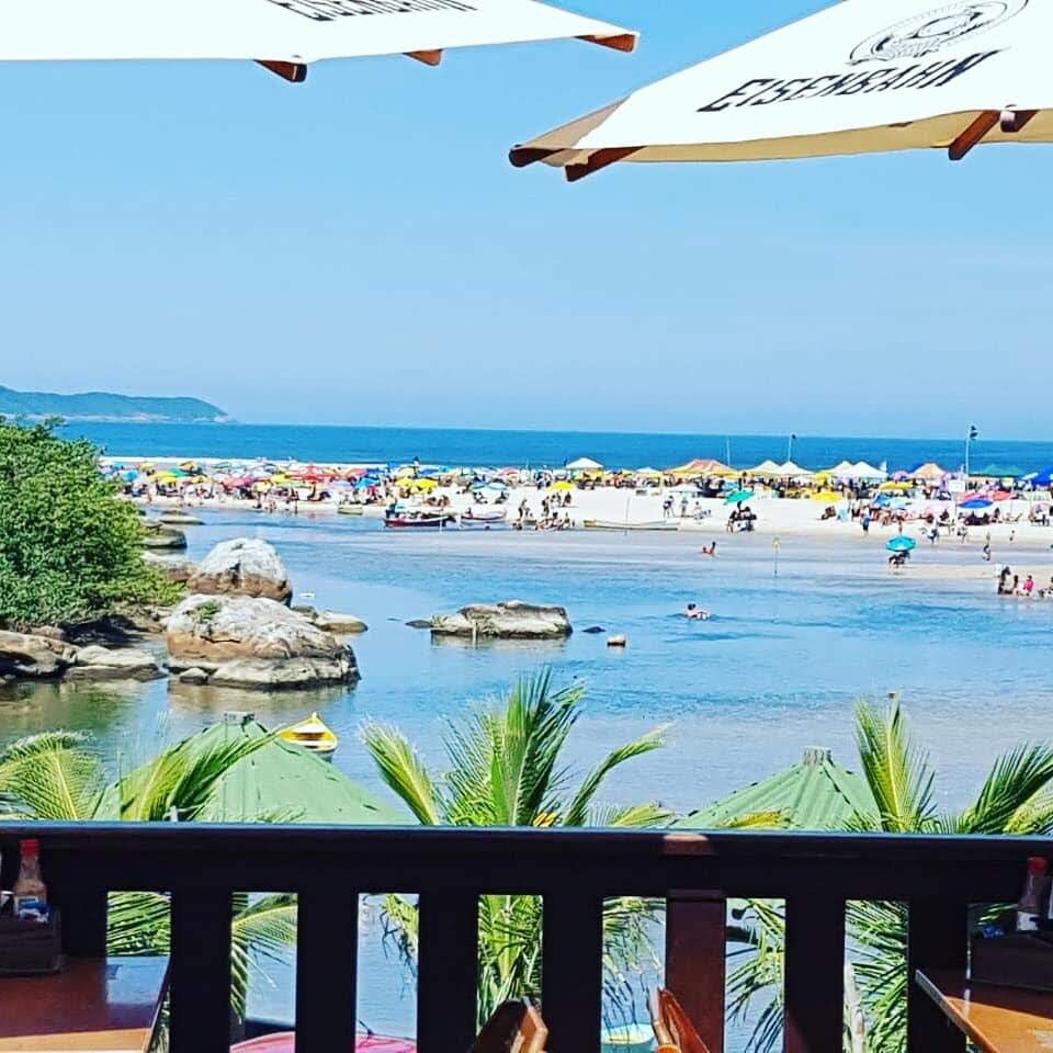 A view overlooking a sandy beach with colorful umbrellas and beachgoers enjoying the sun. Clear blue water reflects the sky, bordered by green vegetation. Large rocks are visible in the water, providing natural accents to the landscape.