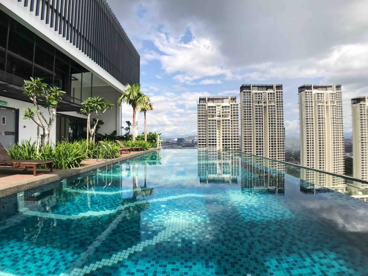An expansive rooftop swimming pool is showcased, reflecting the sky and surrounding cityscape. Lush greenery borders the pool area, offering a serene escape. The modern building structure is visible in the background, with tall buildings complementing the urban skyline.