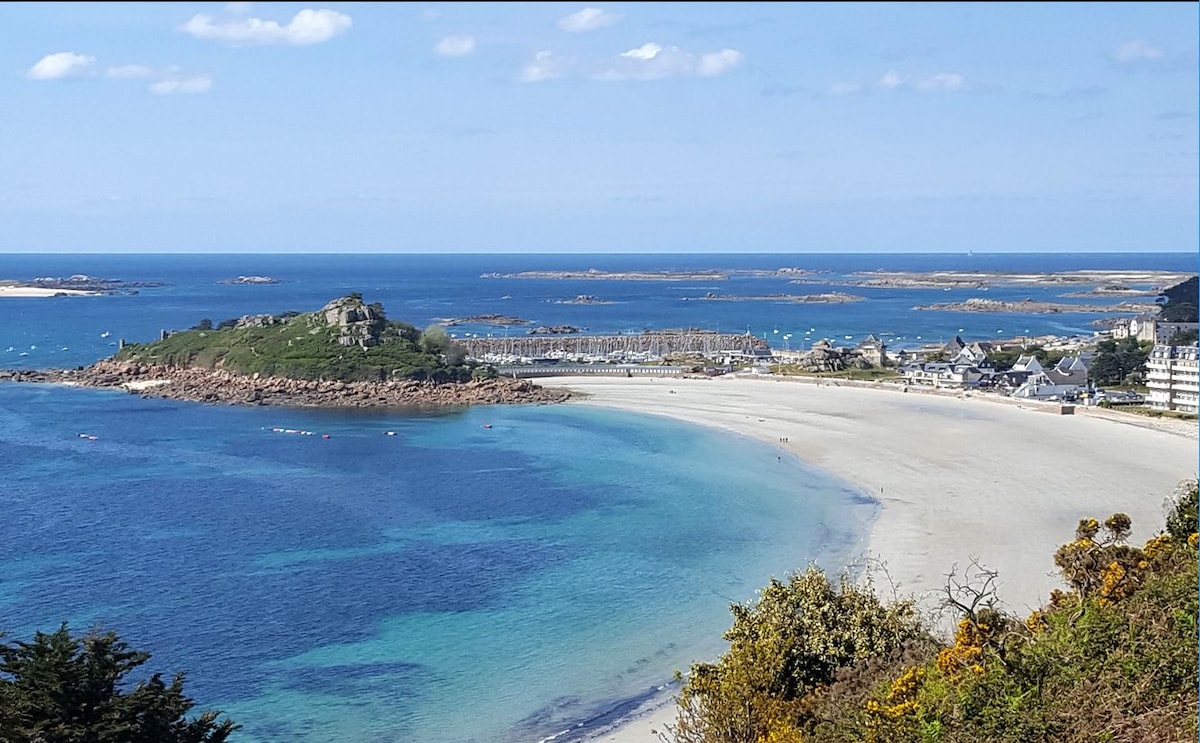 A scenic view captures the beach and coastal landscape, showcasing soft sandy shores meeting the blue waters. Gentle waves lap at the shoreline, while distant islands dot the horizon. The sky is clear, with a few wispy clouds, highlighting the peaceful seaside environment.
