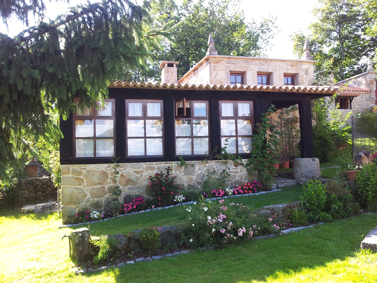 A traditional stone house is framed by lush greenery and colorful flowerbeds. The house features large windows and a tiled roof, allowing natural light to fill the interior. Decorative plants are positioned near the entrance, enhancing the inviting exterior.