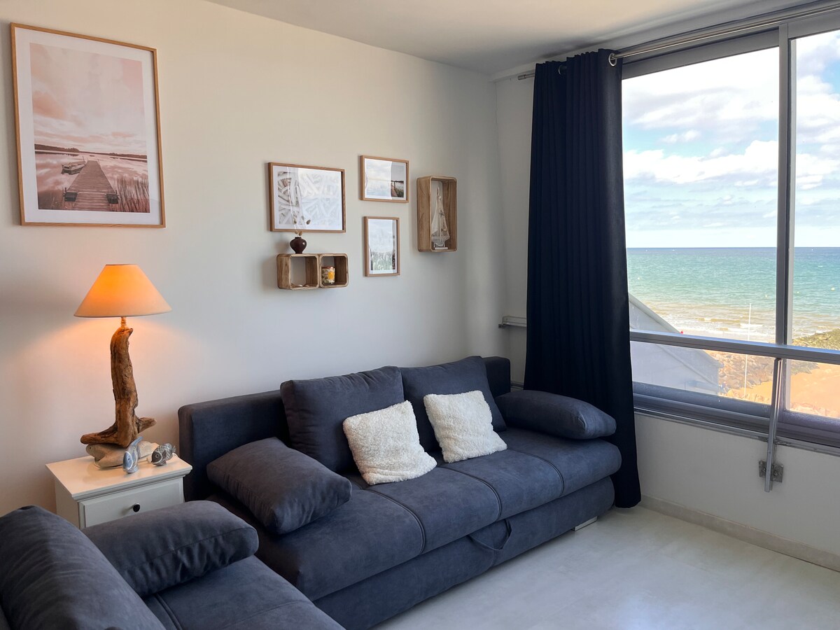 A cozy living room area features a gray sofa adorned with soft cushions. The space is illuminated by a warm lamp, and decorative wall art adds character. Large windows offer a view of the sea, allowing natural light to fill the room.