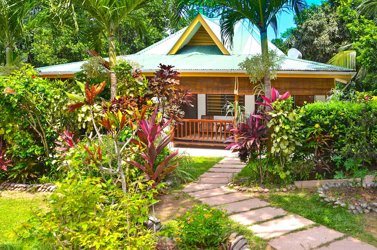 The exterior of the bungalow is framed by lush tropical gardens, showcasing a variety of vibrant plants and flowers. A stone pathway leads to a covered front porch, which features wooden railings. A distinctive peaked roof adds character to the structure, highlighting its harmonious connection to nature.
