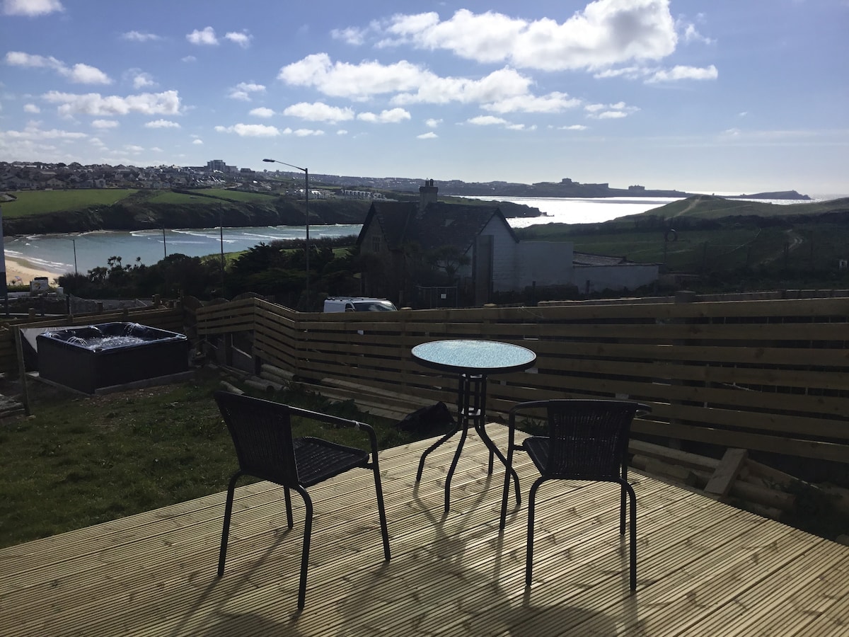 A private decking area features a round table and two chairs, overlooking scenic coastal views under a clear sky. In the foreground, a hot tub is positioned to the left, surrounded by grass and wooden fencing.