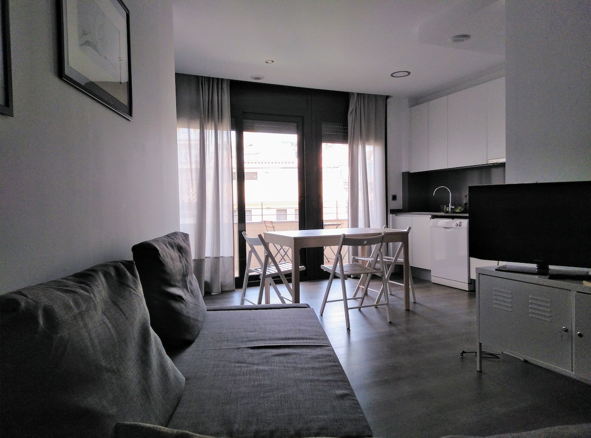 A bright living space is shown, featuring a comfortable sofa and a dining area with a small table and two chairs. The modern kitchen is visible in the background, equipped with white cabinetry and appliances. Natural light enters through large windows with sheer curtains.