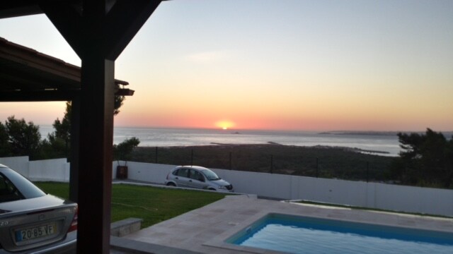 A serene sunset is captured over the horizon, with soft hues of orange and pink reflecting on the water. A swimming pool sits in the foreground, while cars are parked nearby on a neatly kept lawn, surrounded by a white fence and trees.