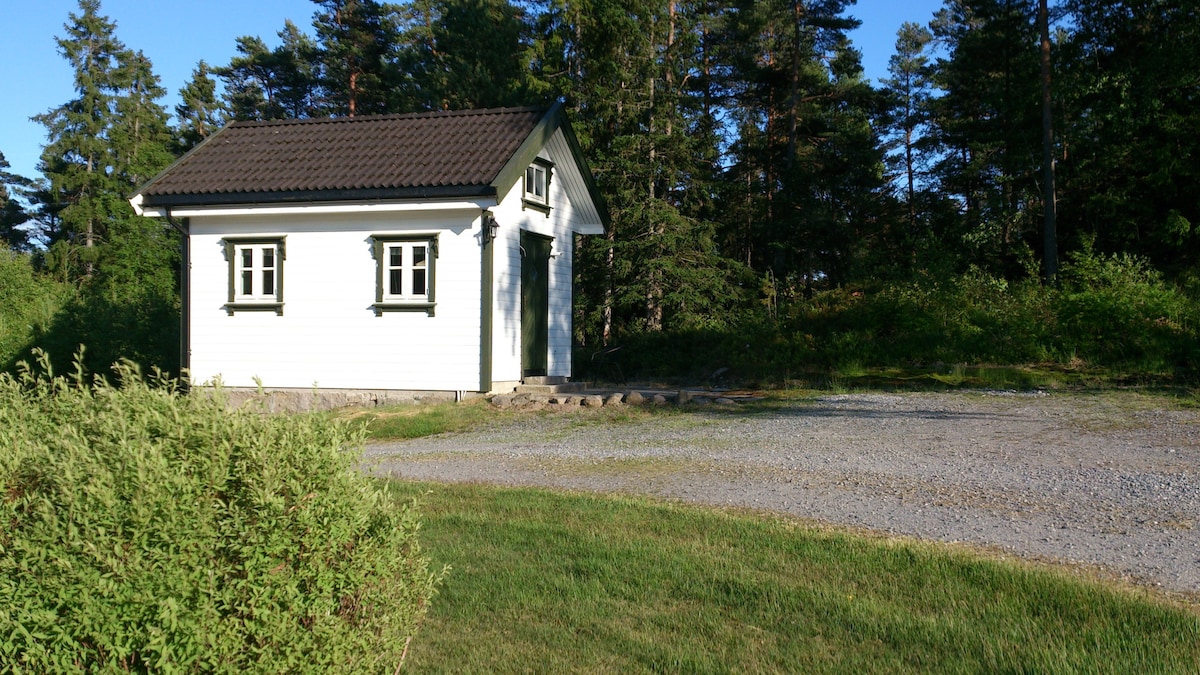 A small, white guesthouse with green accents is surrounded by lush greenery and tall trees. The building features a gabled roof and multiple windows, reflecting a simple architectural style. A gravel pathway leads to the entrance, while a grassy area provides space for outdoor activities.