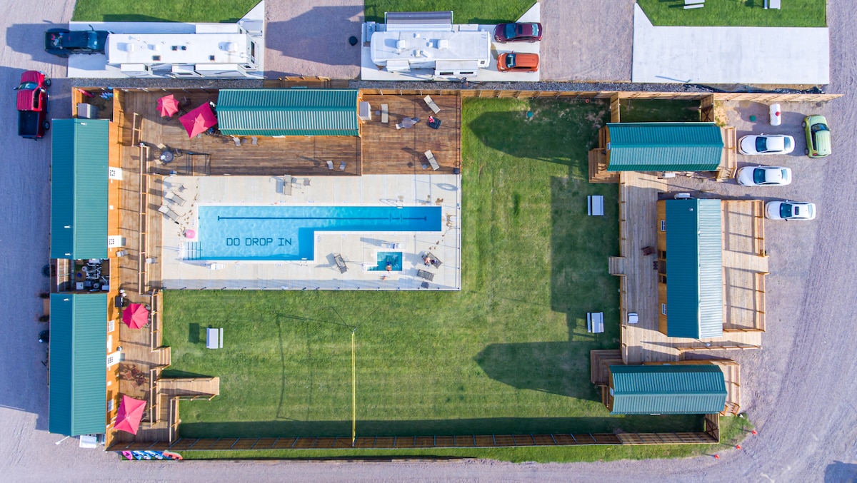 An aerial view captures a sprawling RV resort layout, featuring a rectangular pool surrounded by sun loungers and shaded seating areas. Several RVs are parked alongside, while green spaces enhance the outdoor environment, creating areas for relaxation and recreation.