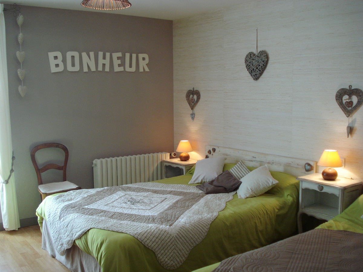 A comfortable sleeping area is furnished with two spacious beds, each adorned with soft pillows and a textured blanket. Warm table lamps sit on nightstands, providing a gentle glow. Decorative hearts and the word 'BONHEUR' adorn the walls, creating a serene atmosphere.