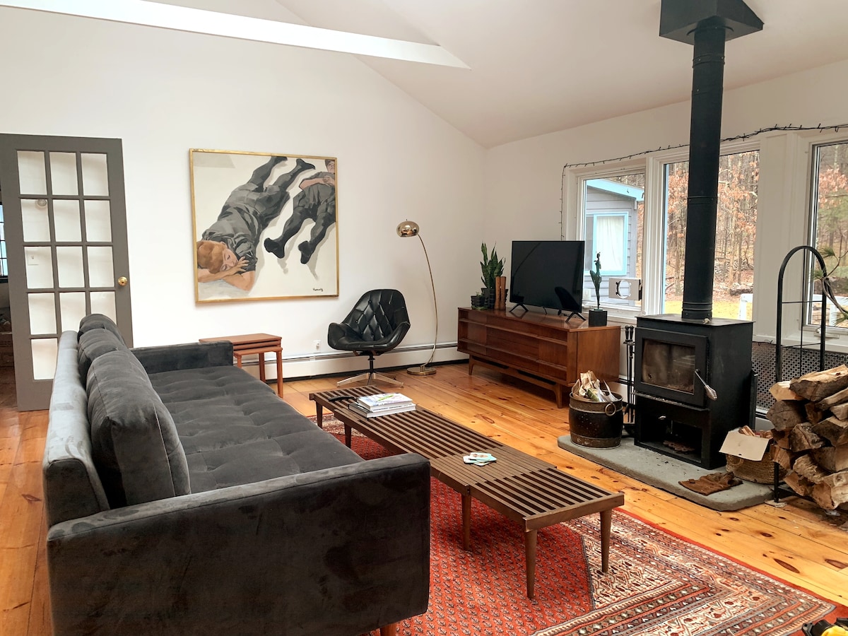 A spacious living area features a charcoal sofa paired with a sleek black armchair. Natural light streams through large windows, illuminating an art piece above a wooden coffee table. A wood-burning stove adds warmth, complemented by a stack of logs nearby.