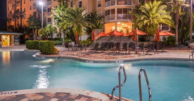 A heated outdoor pool is framed by palm trees and lounge chairs, creating a relaxing atmosphere. The pool deck features inviting umbrellas and landscaping that enhances the area. A well-lit building with large windows is visible in the background, adding to the resort’s welcoming environment.
