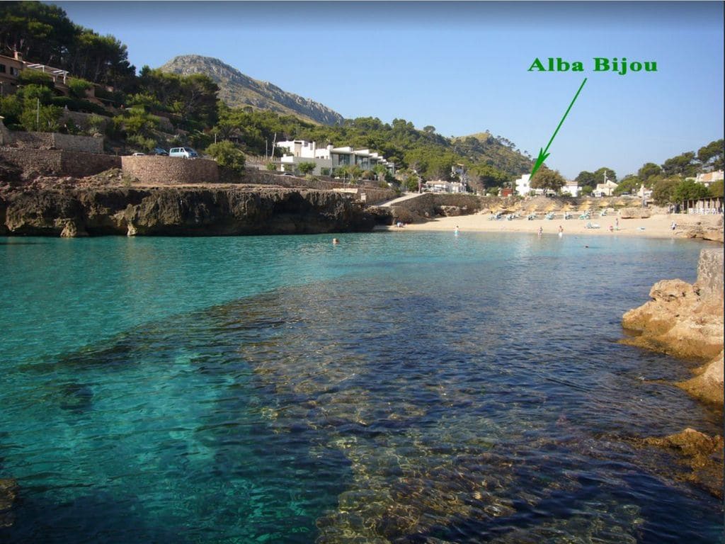 A crystal-clear bay is depicted, with the view extending towards a calm, turquoise water. The shoreline features sandy areas populated with beachgoers, while rocky formations frame the scene. In the background, the Alba Bijou is marked, nestled amidst trees and buildings on the hillside.