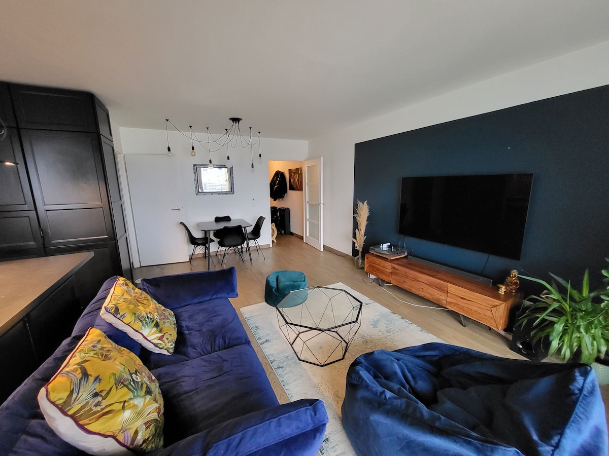 A modern living area features a dark blue accent wall complemented by a light-toned wooden floor. A large flat-screen television is mounted on a wooden console. A geometric coffee table is surrounded by comfortable seating options, including two bean bags and a sofa adorned with colorful cushions.