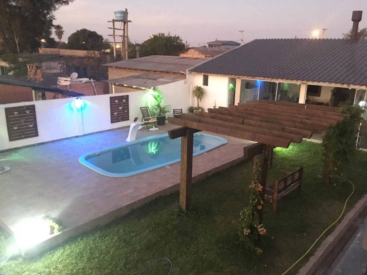 An outdoor area features a rectangular swimming pool surrounded by a well-maintained lawn. A pergola provides shade over seating, while subtle lighting enhances the space. The background includes a house with large glass doors, contributing to a relaxed atmosphere for guests.