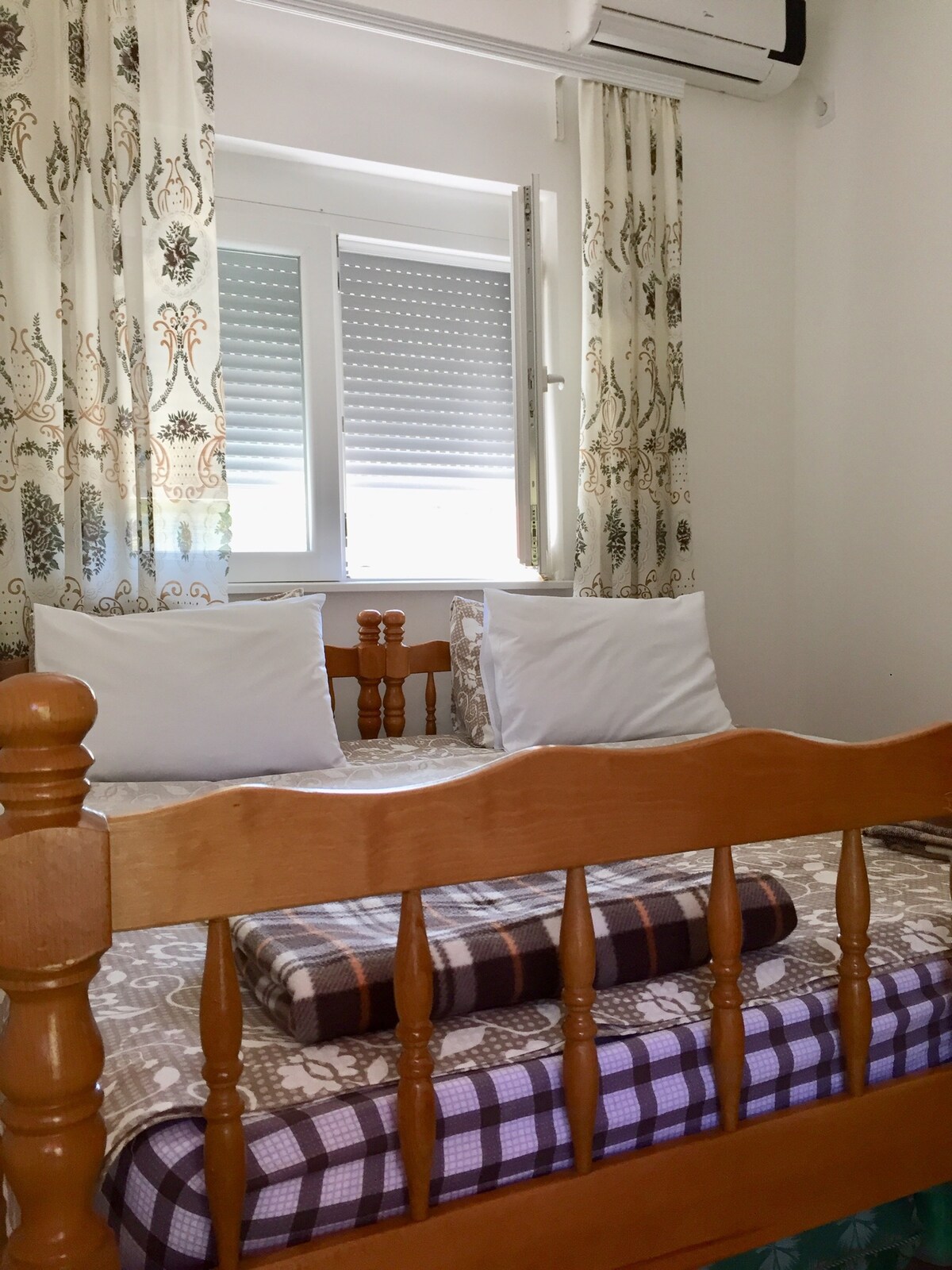A bedroom is presented with a wooden bed featuring a patterned mattress and additional throw pillows. Light enters through a window covered with sheer curtains, allowing for a bright and airy appearance. Air conditioning is visible above, ensuring a comfortable climate.
